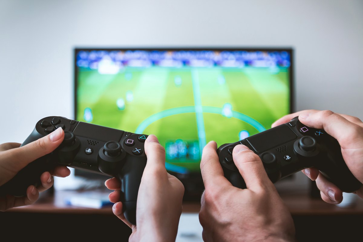 Two hands holding game controllers in front of a TV